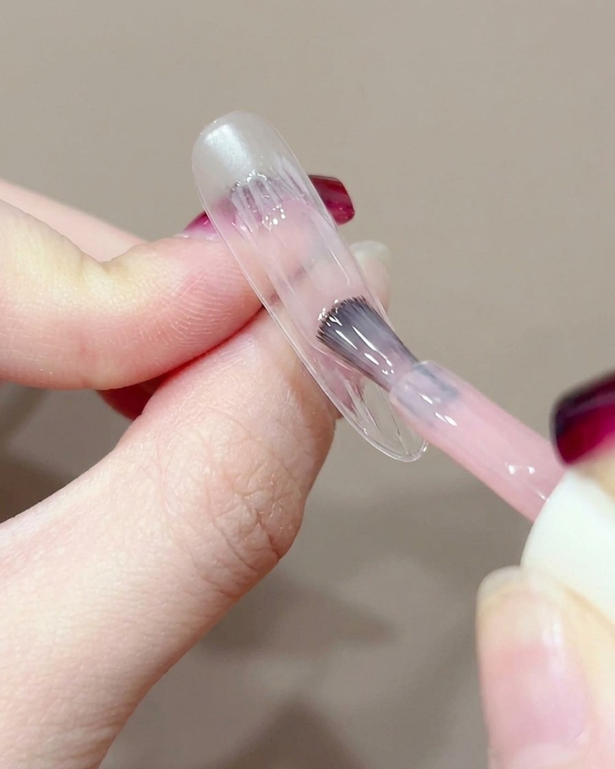 A close-up of a gel brush applying a small dab of peach gel to the center of a bare nail.