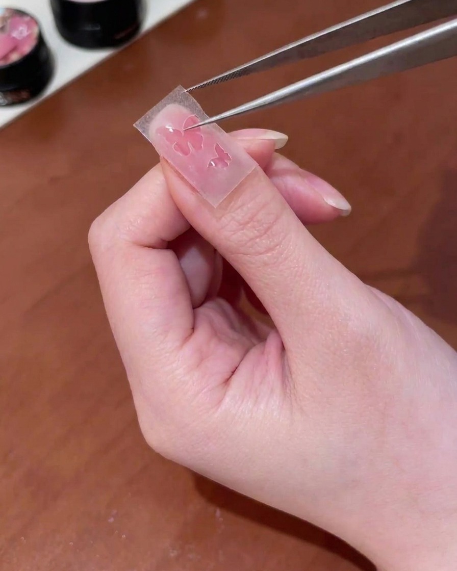 Tweezers placing a butterfly-shaped cutout sticker onto a nail surface.