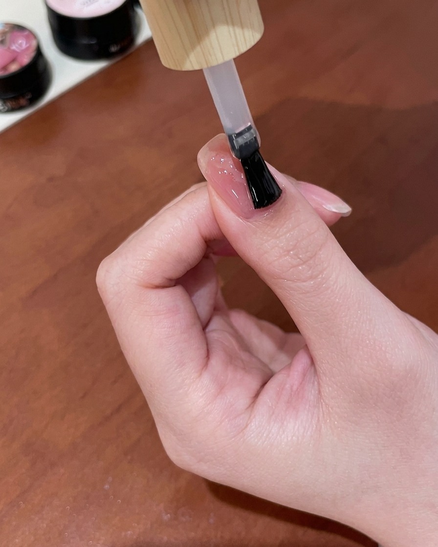 A brush applying a layer of clear, shiny gel top coat over a finished nude pink nail featuring a gold cat-eye butterfly design.