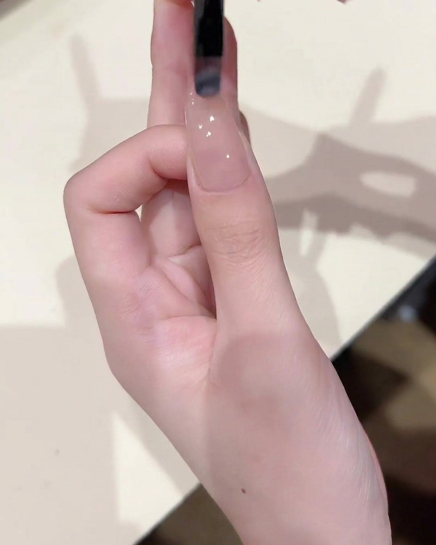A close-up of a nail brush applying a thin, even coat of sheer nude gel to a nail plate already coated with a base layer.
