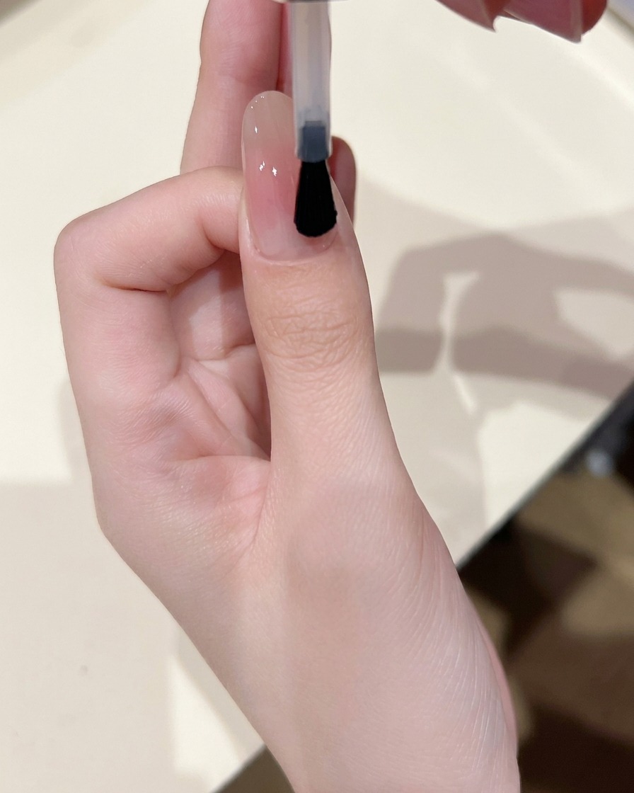 A nail brush applying a thin layer of clear, glossy base gel over a nail that has a soft blush center.
