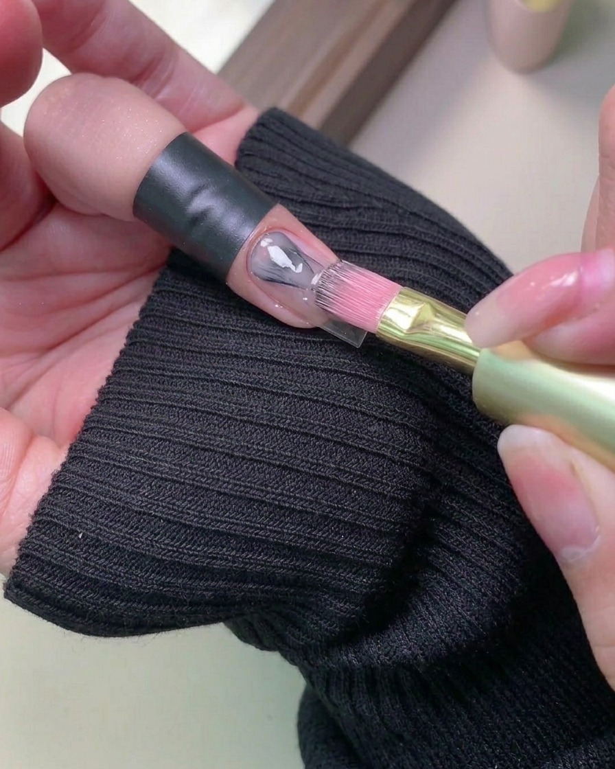 A close-up view of a brush applying a soft, light-colored gel polish onto a natural nail.