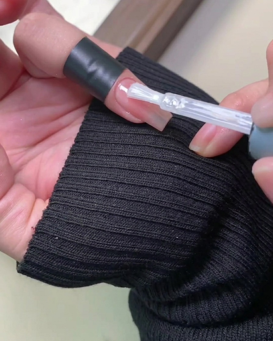 A close-up of a brush applying a clear, glossy non-wipe top coat over a soft, blush-pink gradient nail.