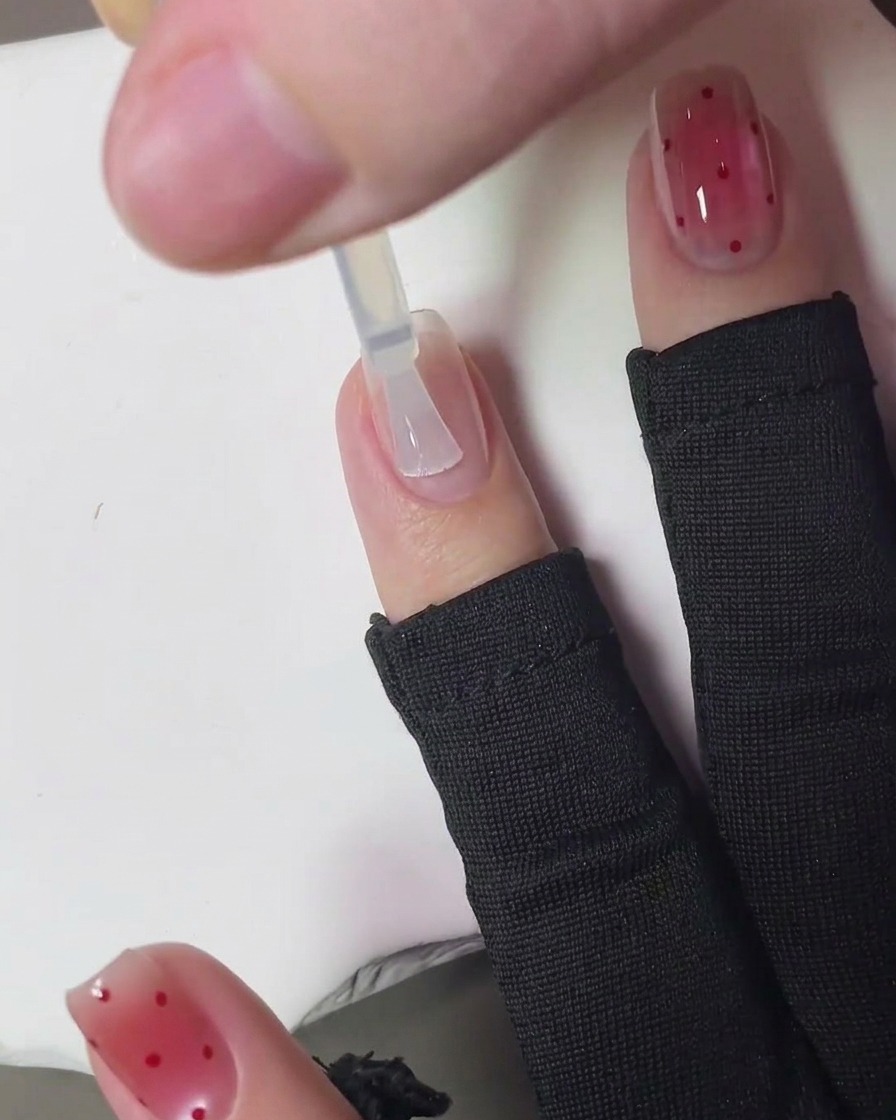 A brush applying a clear, glossy top coat over the finished strawberry-dotted nail art.