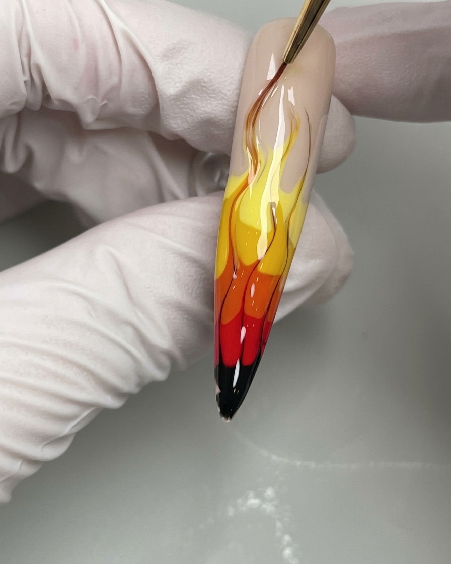 A close-up of a nail brush applying thin black gel lines to define the edges of a yellow, orange, and red flame nail art design.