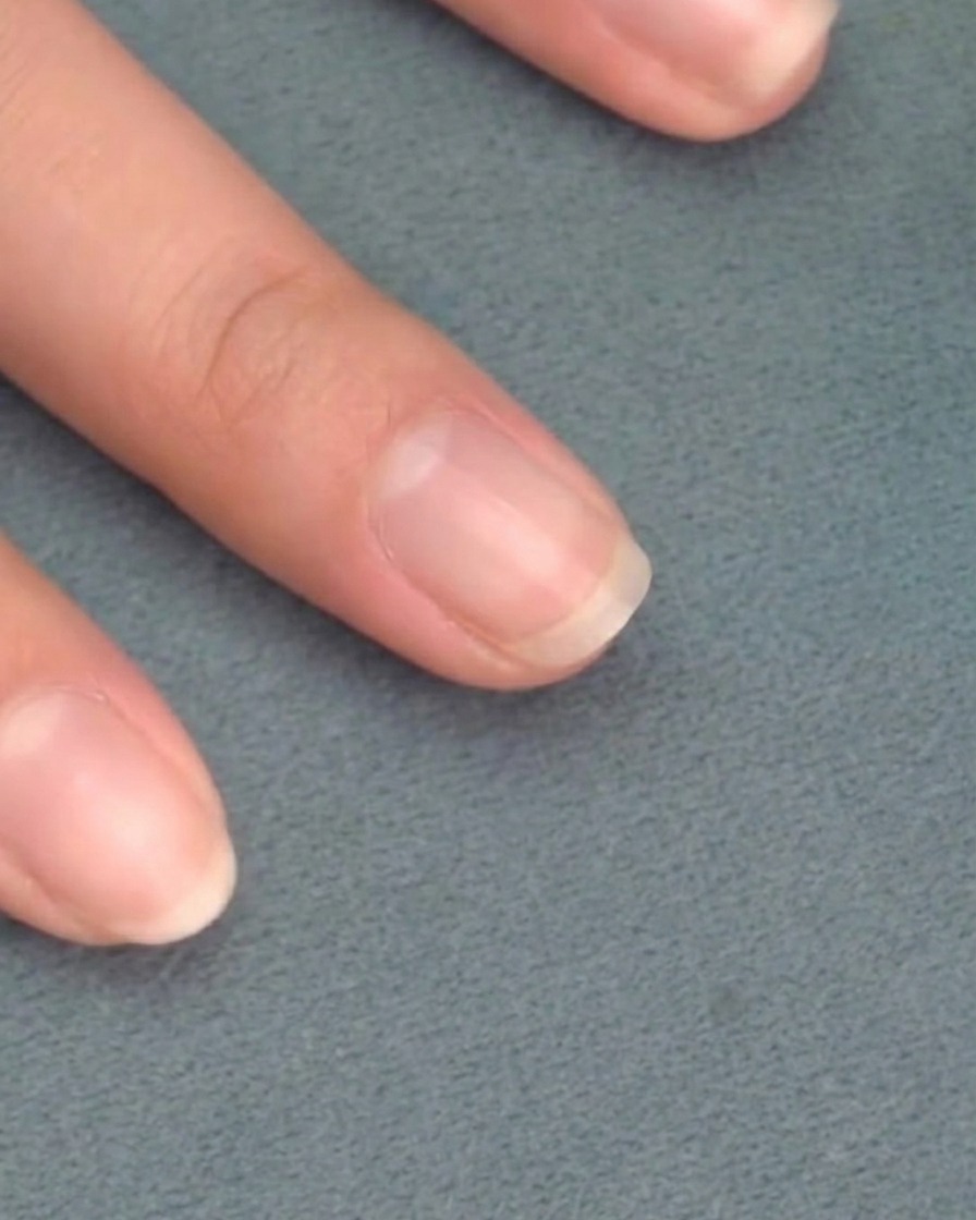 A brush applying a sheer pink gel polish onto a clean, natural fingernail.
