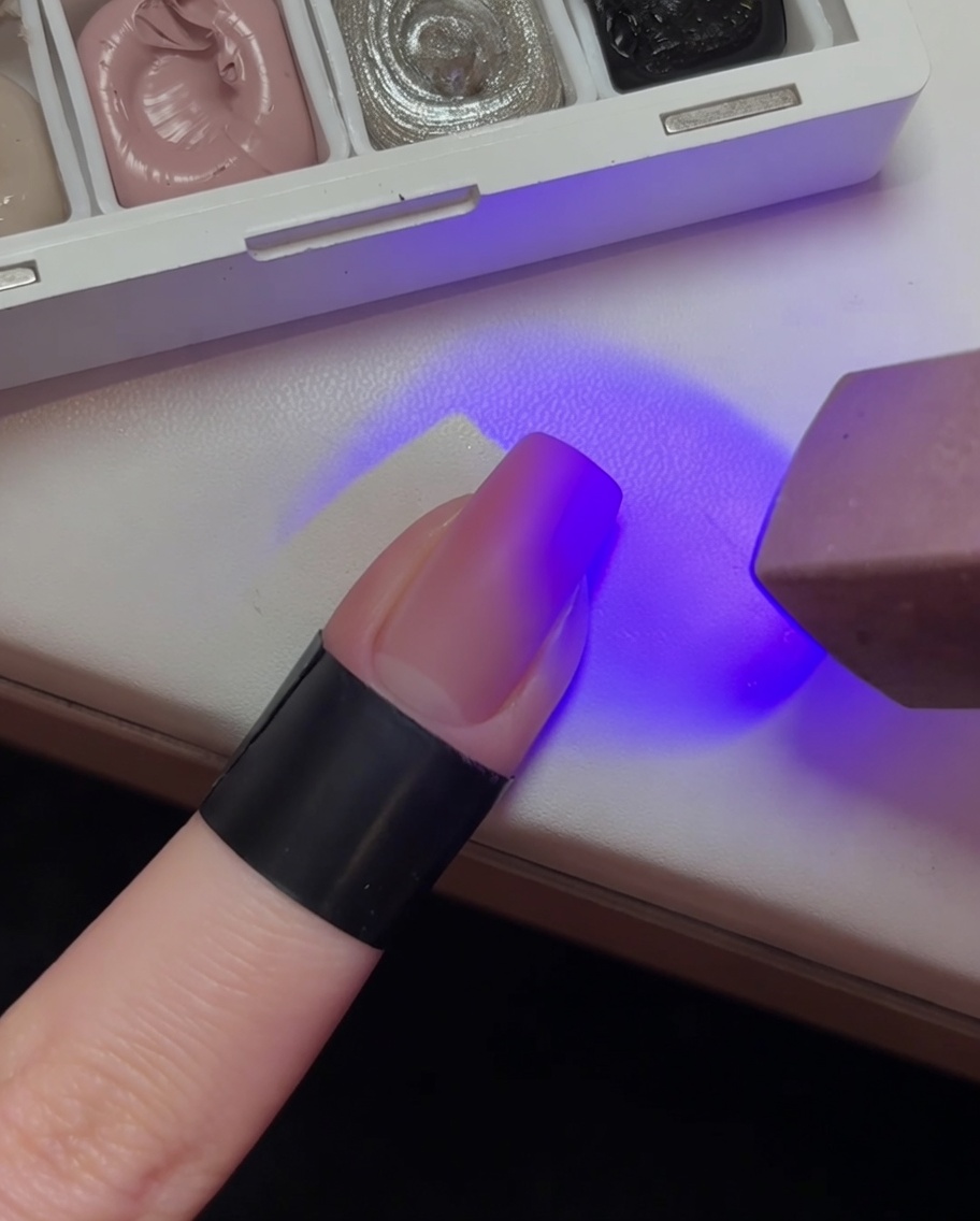 A fingernail with a light pink gradient base being cured under a purple UV lamp light.