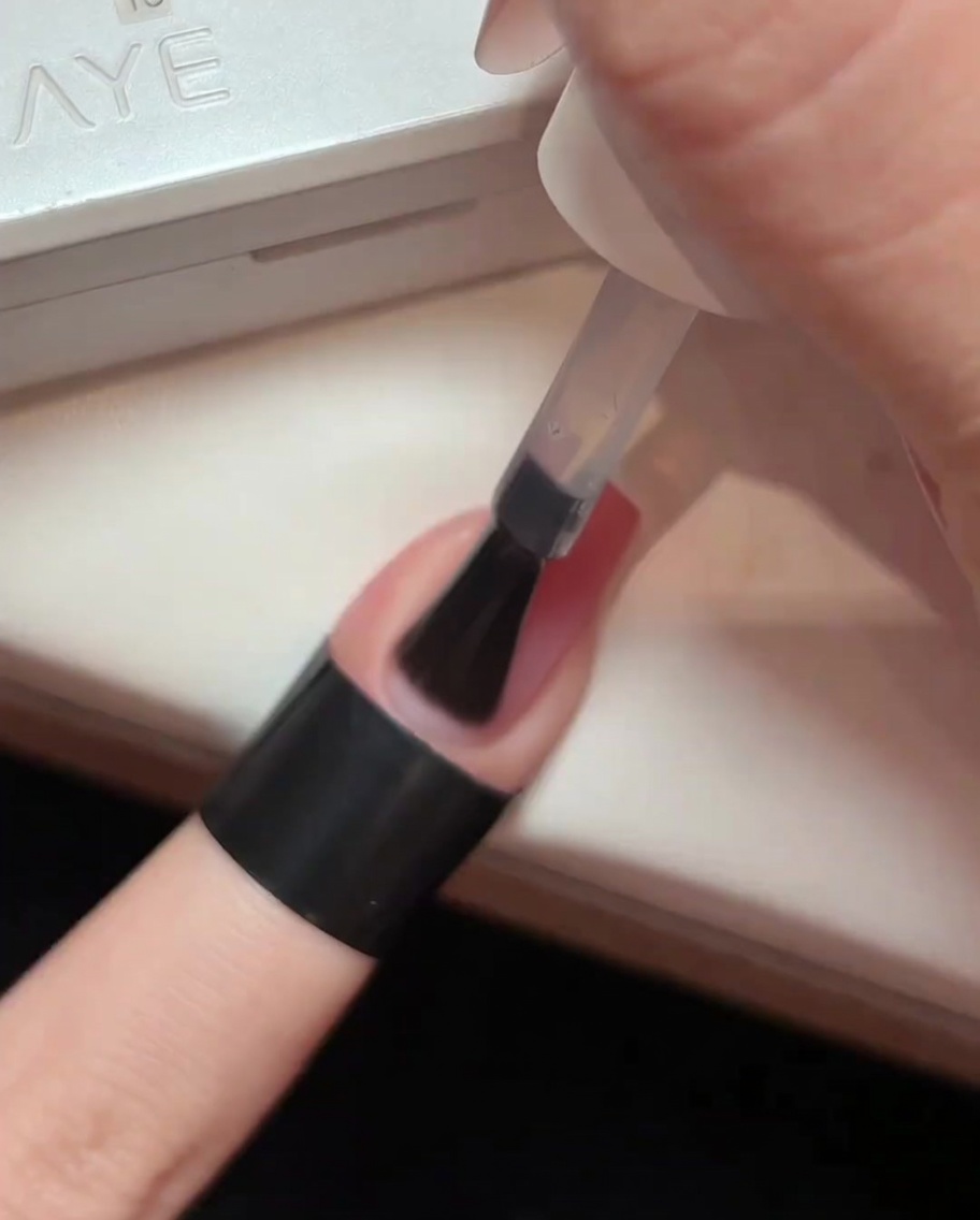 A brush applying a layer of clear glossy top coat over a light pink gradient nail.