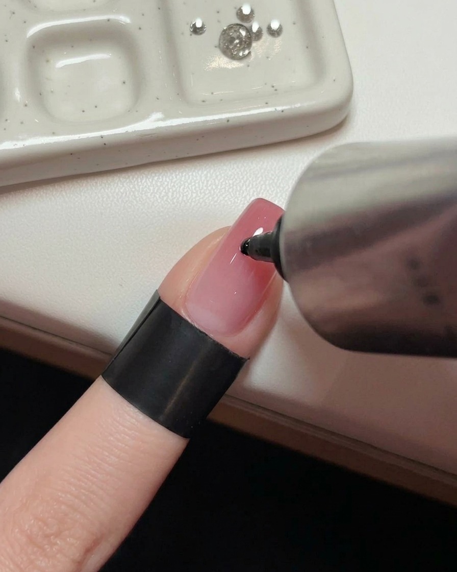 A hand holding a metal tube squeezing a clear drop of adhesive gel onto the center of a glossy pink manicured nail.