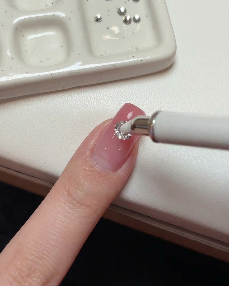 A white wax-tipped pen placing a large round 5mm crystal onto the center of a pink nail.