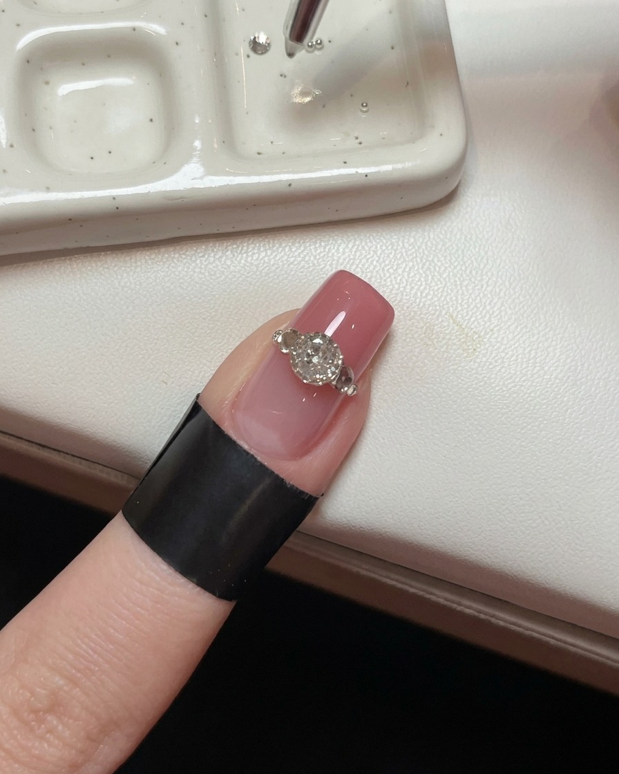A close-up of a nail with a large crystal, showing a precision tool placing tiny steel beads around its base to create a prong effect.