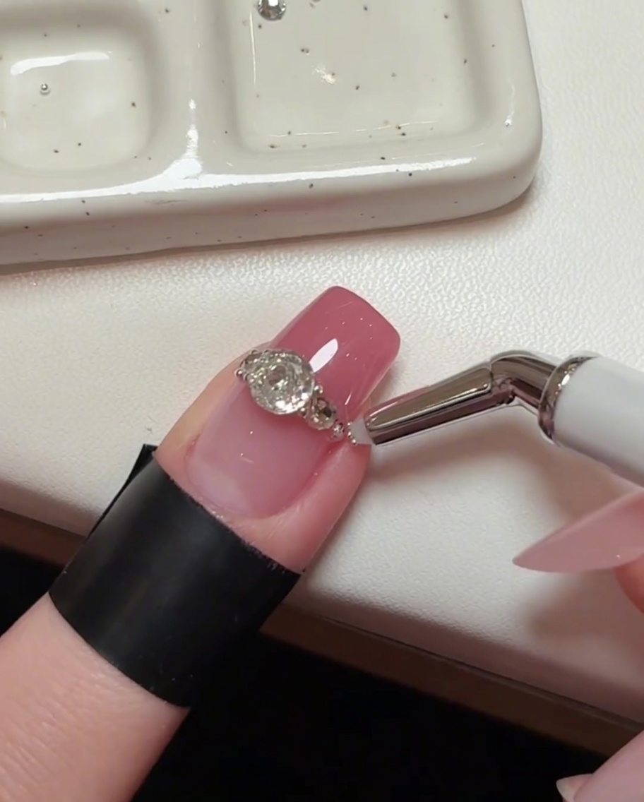 A detailed shot of the nail art, showing a precision tool placing microscopic steel beads into the small spaces between larger crystals.