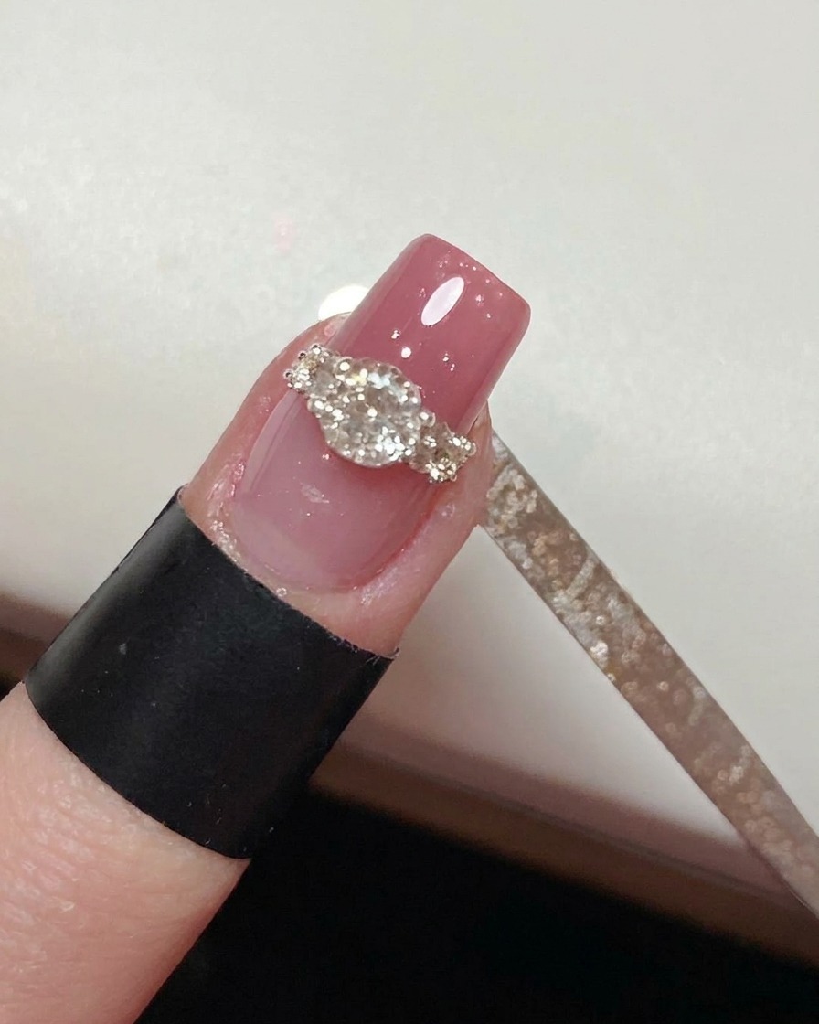 A fine liner brush carefully painting top coat around the base of a completed crystal ring design on a pink nail.