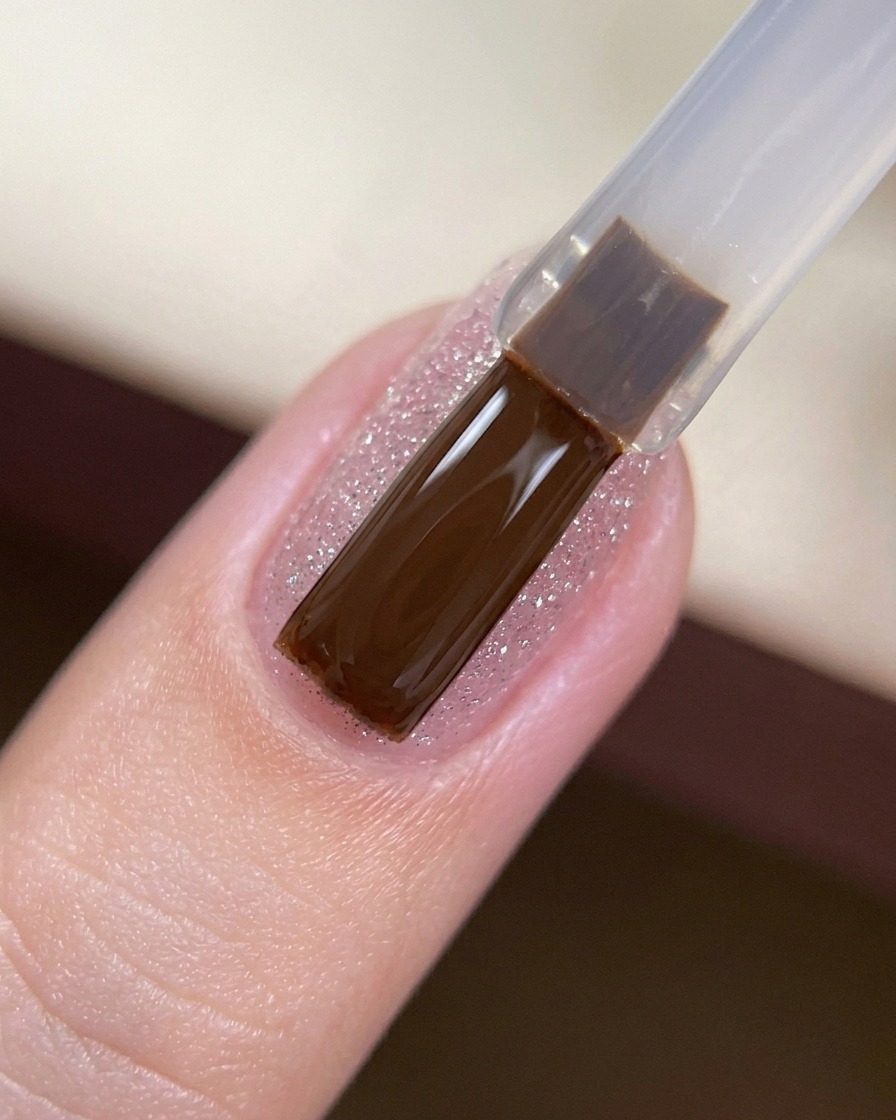 A gel brush applying a clear, glossy top coat over a glitter-covered nail.