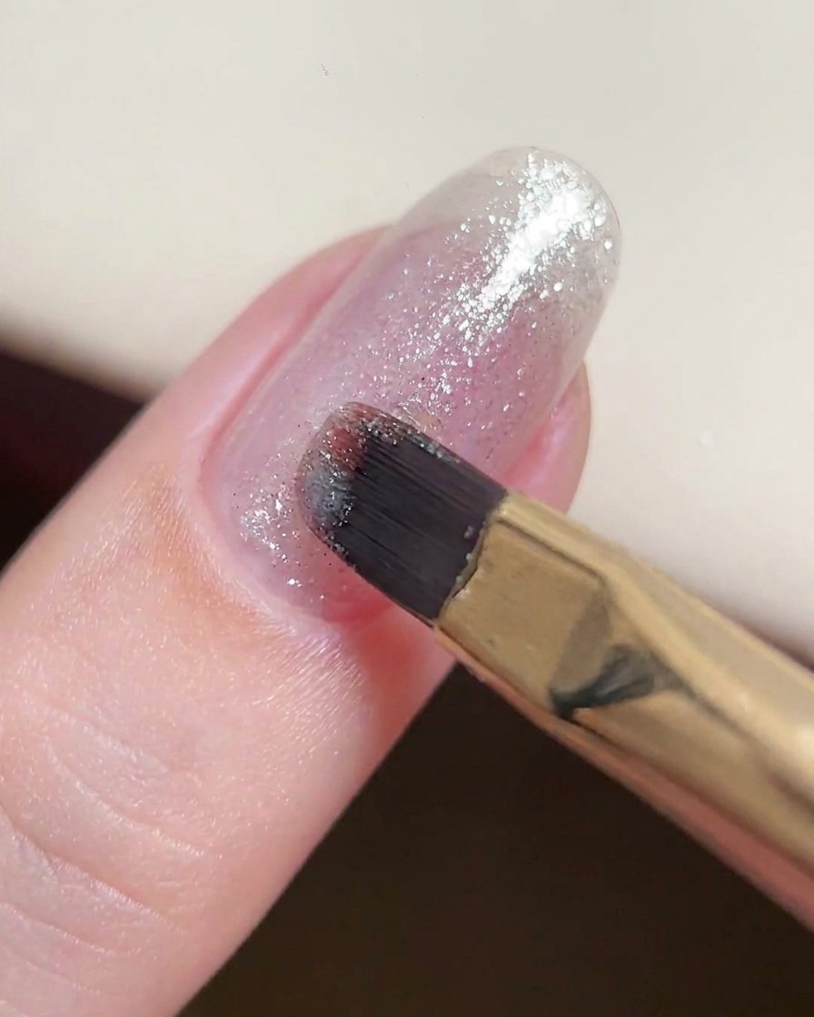 A gel brush applying a thin, consistent layer of fine silver glitter gel onto a natural nail.