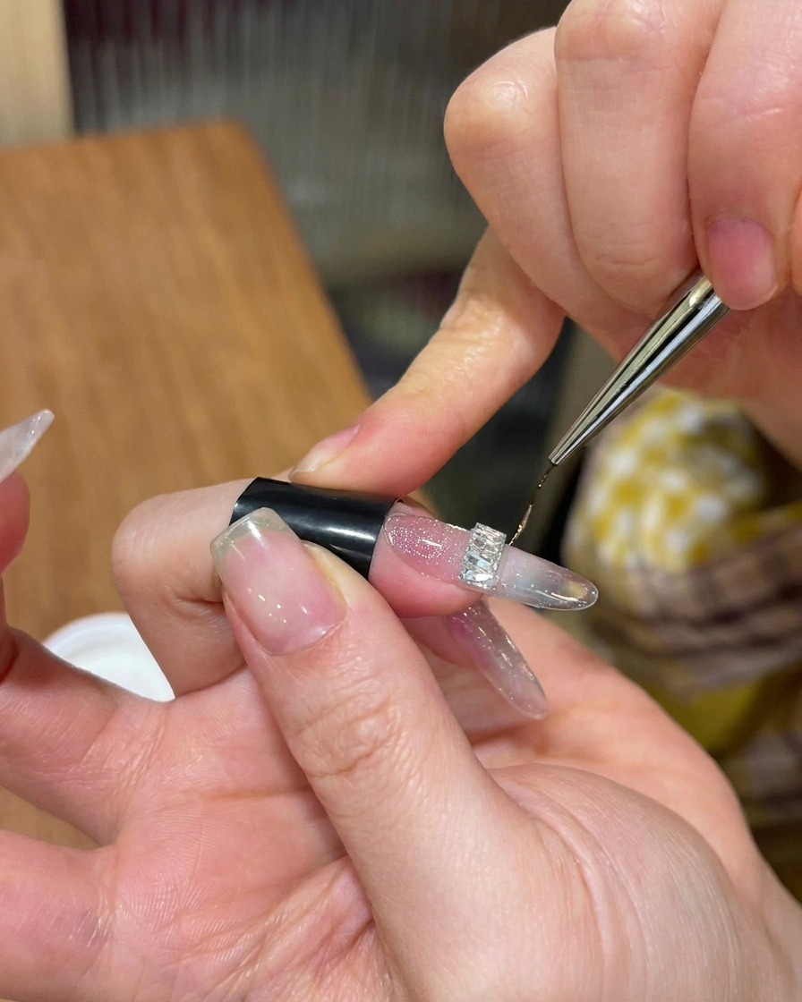 A close-up view of a thin nail art brush applying clear hard top gel around the base of a silver rhinestone charm on a glittery nail.