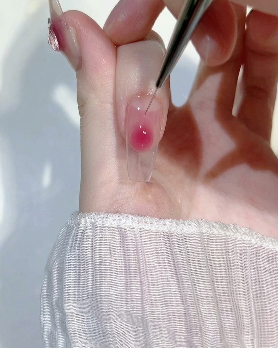 A close-up of a thin liner brush gently blending a drop of pink blush gel onto a clear nail extension to create a soft gradient effect.