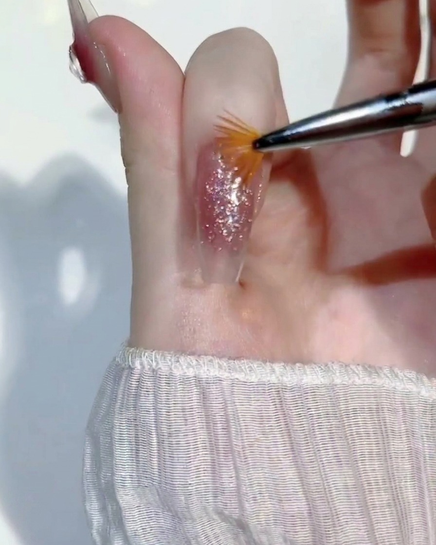 A miniature fan brush being lightly swept across a wet layer of glitter gel on a fingernail to even out the coverage.