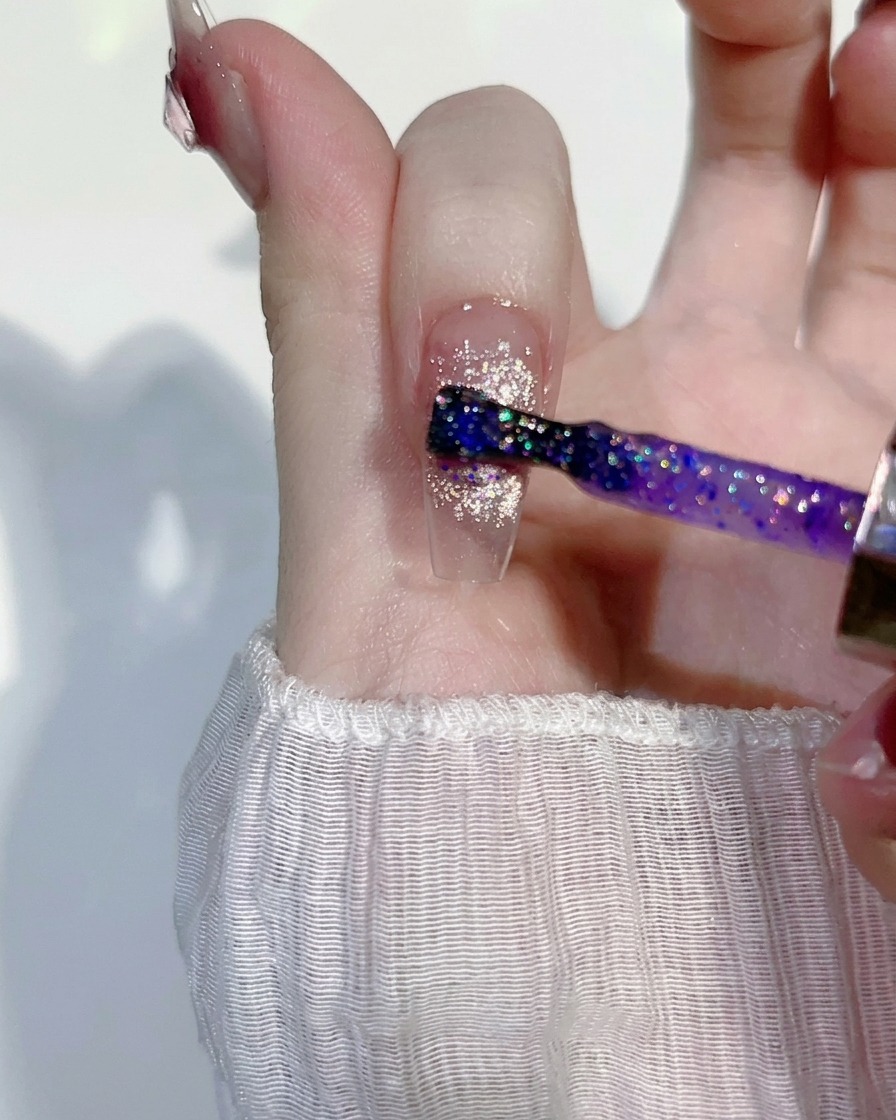 A thin layer of purple-tinted, glittery gel being brushed over an existing pink cat-eye nail design.