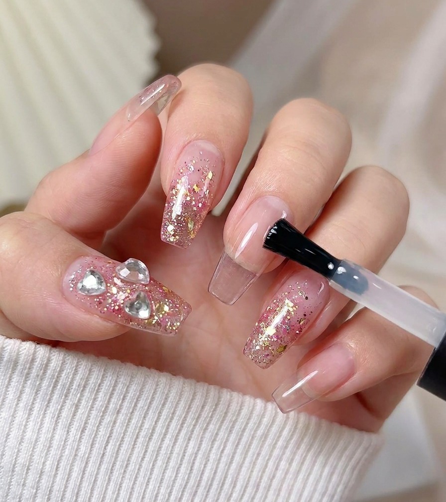 A brush applying a final glossy top coat over a nail decorated with pink glitter and heart crystals.