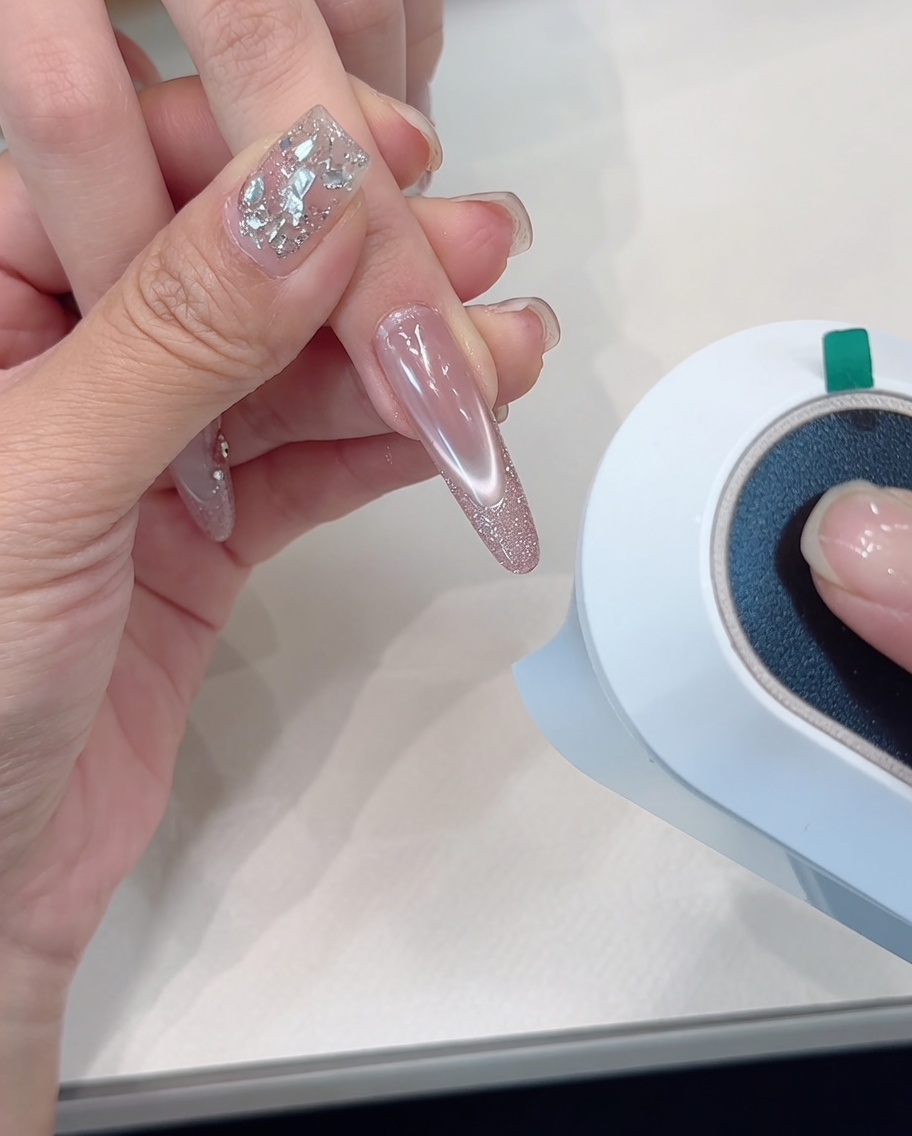 A hand positioned under a nail curing lamp, with the focus on the glitter-tipped nails.