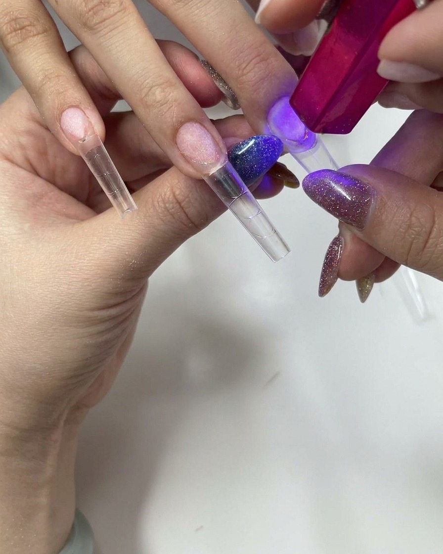 Using a small pink handheld UV/LED lamp to cure a clear press-on nail tip.