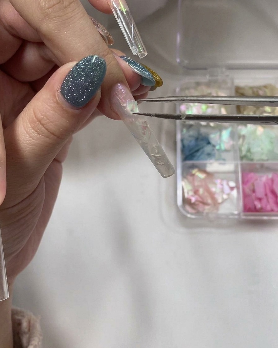 Tweezers carefully placing thin, iridescent shell fragments onto a wet gel layer on a nail tip.