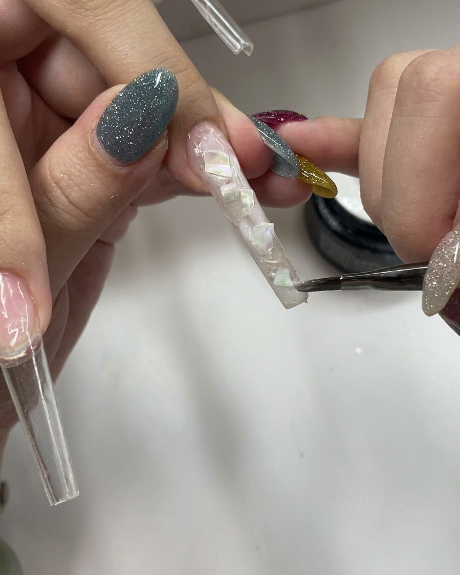 A brush applying a coat of sheer, milky white gel over a textured nail surface containing embedded shell pieces.