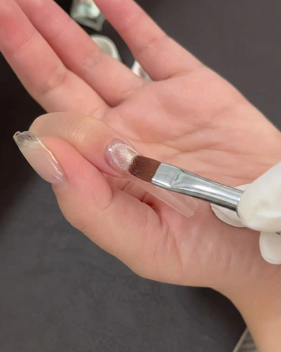 A nail art brush applying a thin layer of shimmering cat-eye gel polish to a clear nail tip.