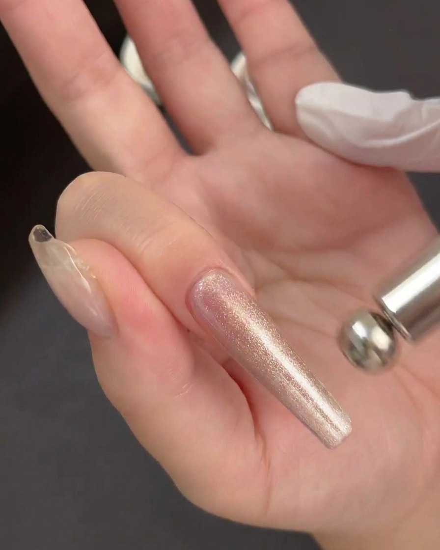 A hand holding a cylindrical magnetic tool hovering just above a wet, sparkly gel application on a clear nail extension.