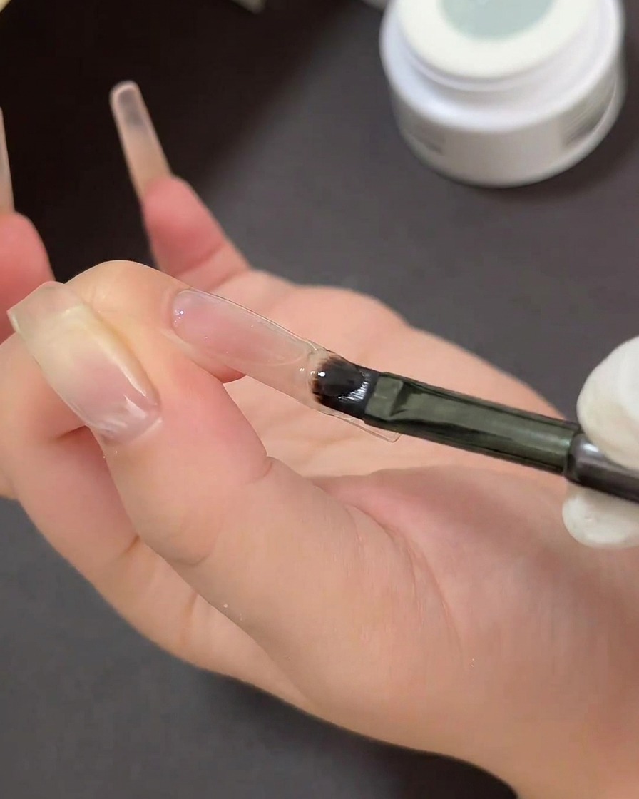 A flat gel brush painting clear sticky gel onto the tip of a clear nail over a curved French tip stencil.