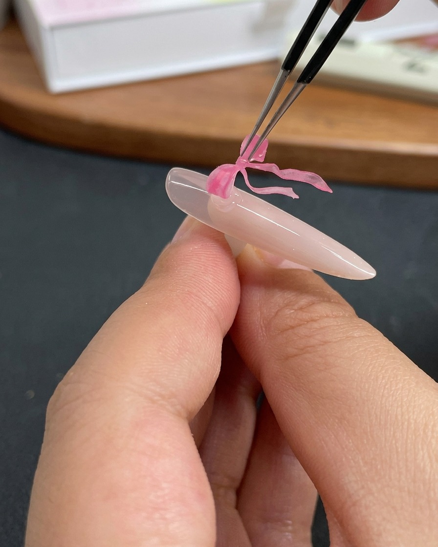 Tweezers placing a delicate pink bow charm onto a fingernail coated in clear gel.