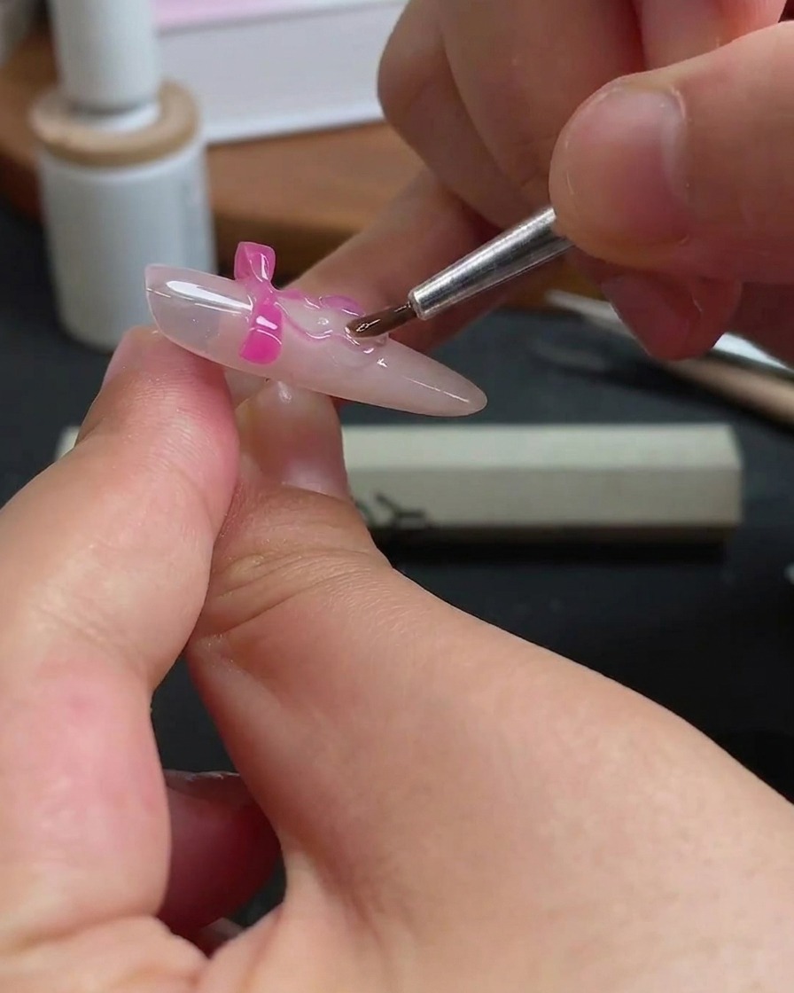 A thin nail brush carefully applying clear gel around the edges of a pink bow charm on a fingernail.