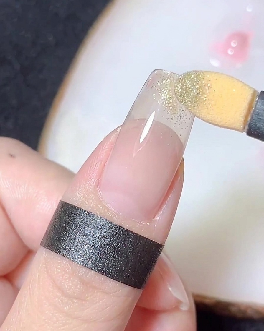 A sponge applicator rubbing gold chrome powder onto the tip of a clear nail tip to create a gradient.
