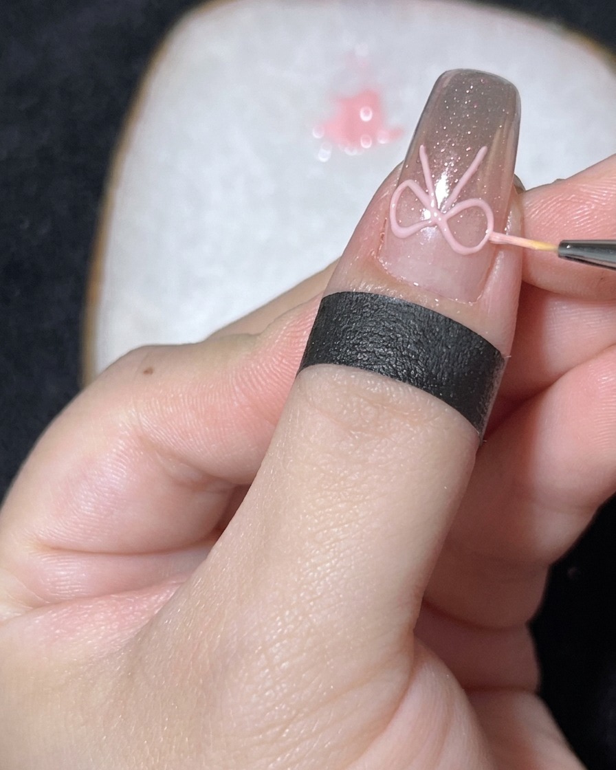 A close-up of a fine brush placing a small, white pearl in the center of a 3D gel bow on a chrome nail.