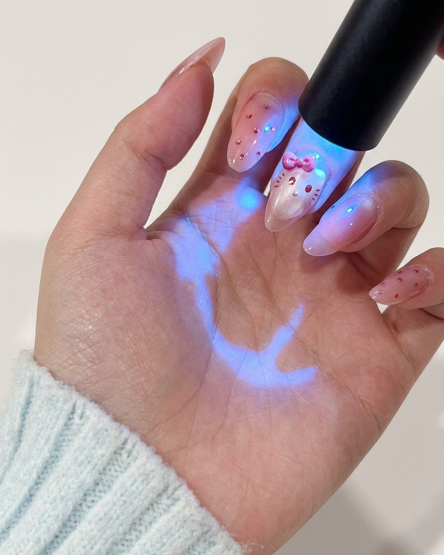 A hand holding a UV lamp over the fingernail to cure the red painted character design.