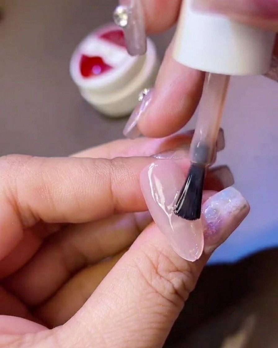 A hand holding a polish brush, applying a layer of matte top coat over a translucent nail tip.