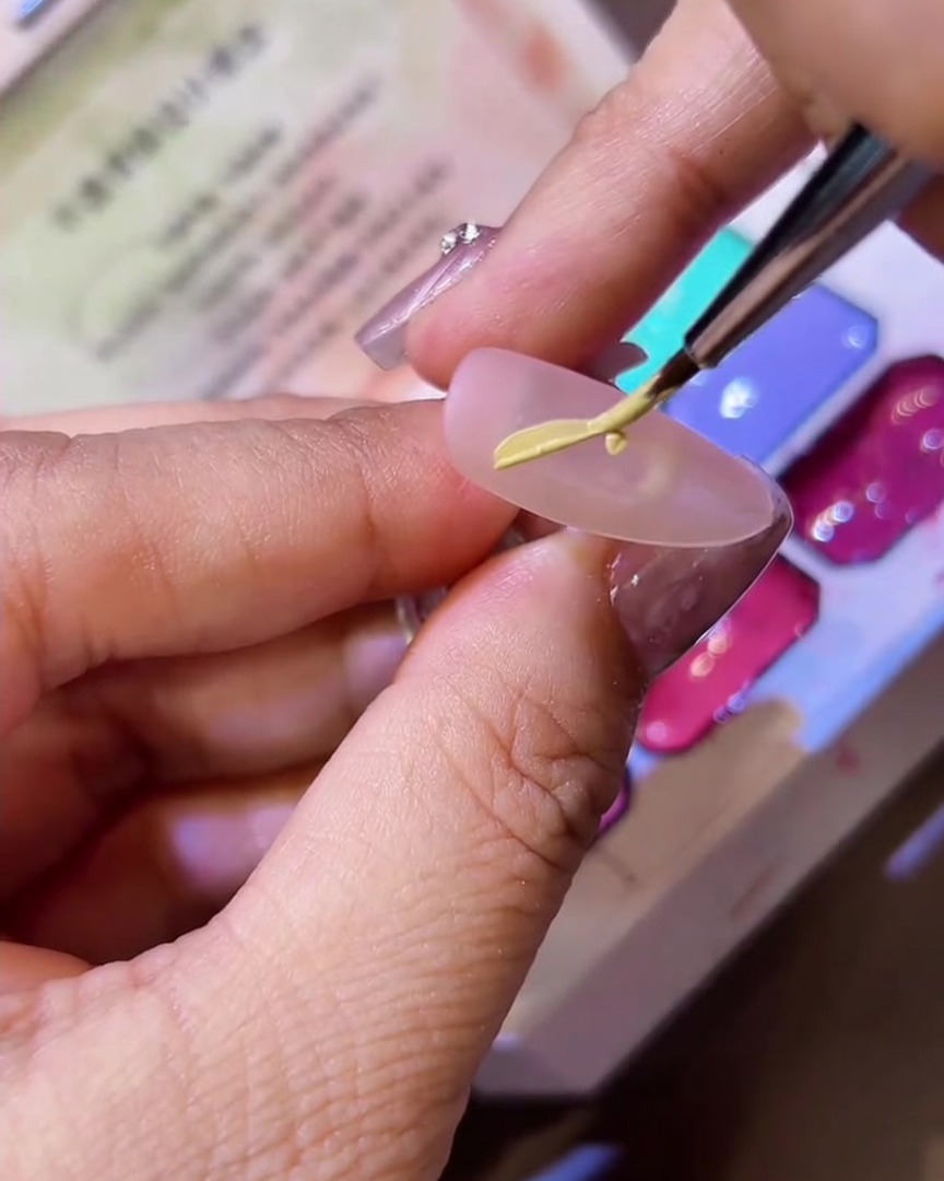 A fine liner brush drawing the shape of half a butterfly wing onto a matte nail tip using yellow gel polish.