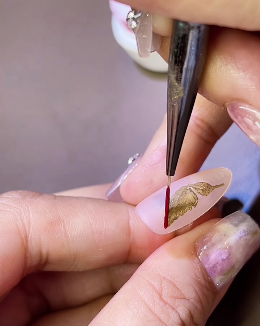 A fine liner brush applying a thin red gel outline around a gold metallic butterfly wing on a nail.