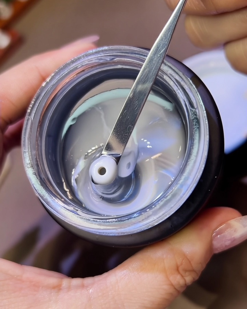 A close-up view of a metal tool placing small beads of thick, clear sculpting gel onto a nail.