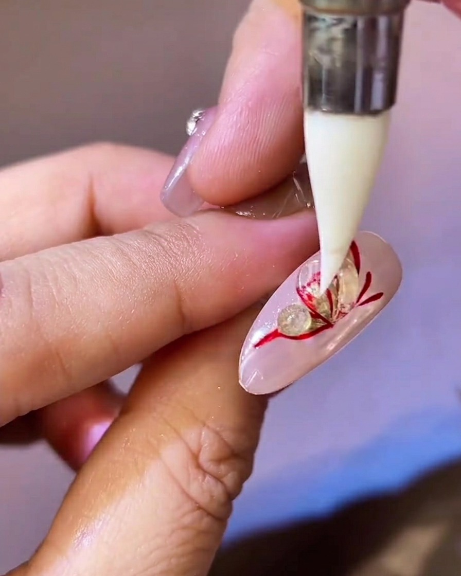 A pointed silicone tool pressing and flattening a bead of clear sculpting gel over a red and gold butterfly wing on a nail.