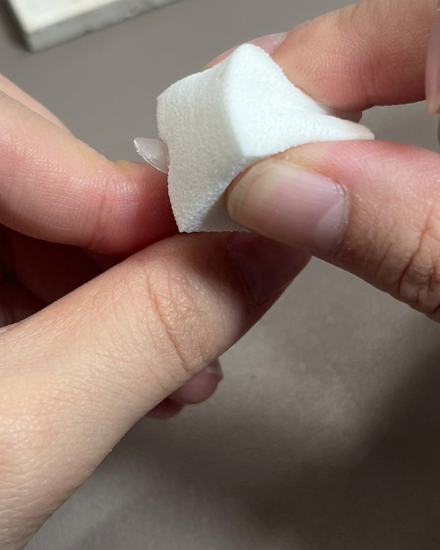 A close-up of a brush applying a sheer pink gel polish onto a nail tip.