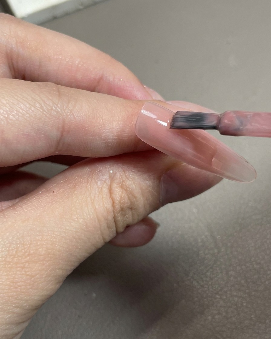 A close-up of a brush applying sheer pink gel polish onto a clear nail tip.