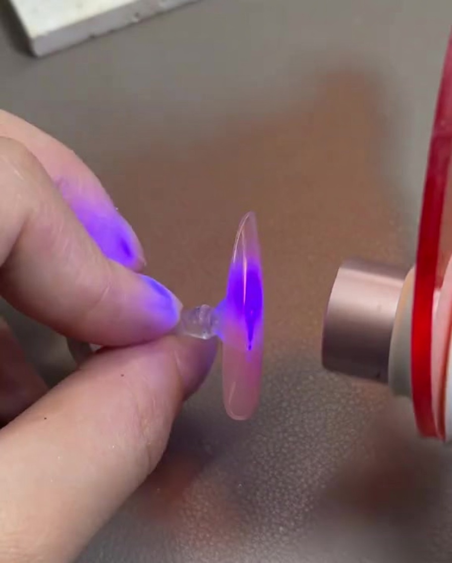 A hand holding a clear nail tip with pink gel under a mini UV/LED curing lamp.