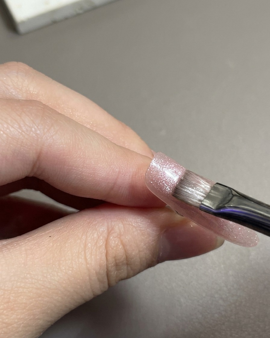 A flat nail brush spreading shimmery pink magnetic cat-eye gel over the nail tip.