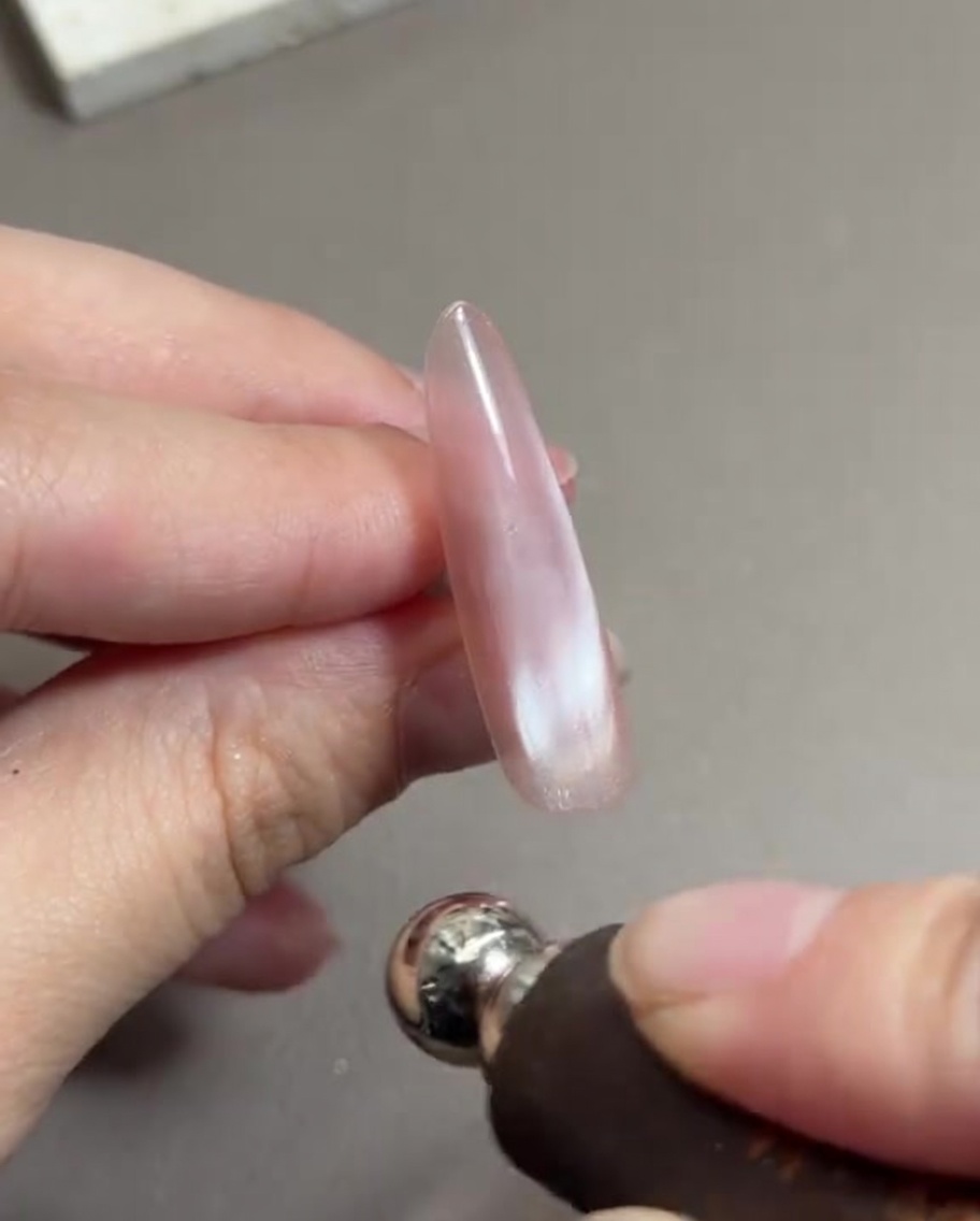 A hand holding a cylindrical magnet tool close to a long fingernail painted with sheer pink wet cat-eye gel, shifting the magnetic particles.