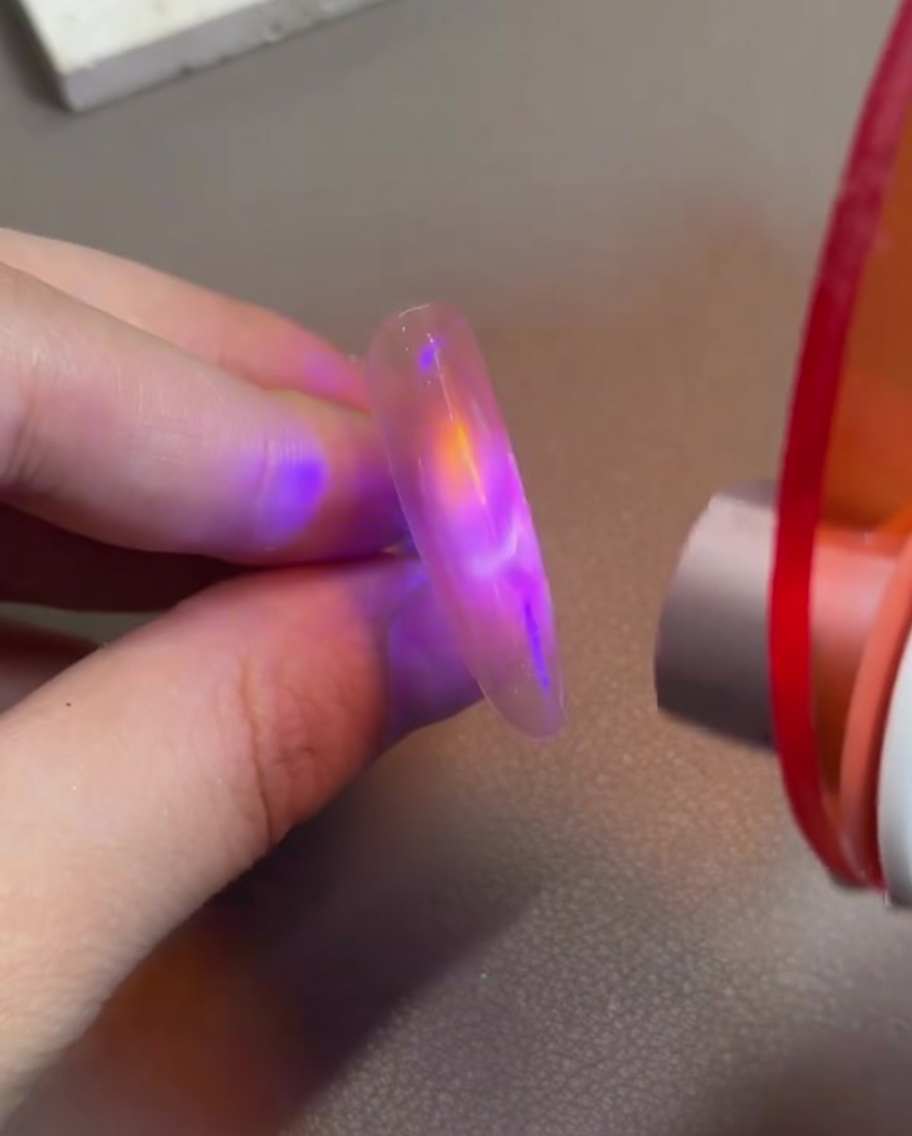 A finger with pink cat-eye gel being held under a small handheld LED curing lamp to set the magnetic design.