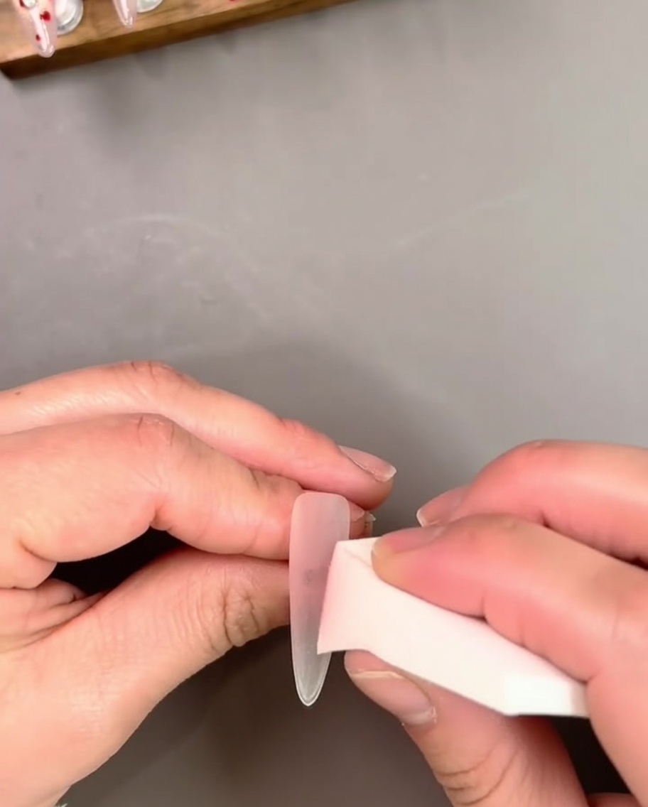 A hand holding a nail extension while applying a sheer pink gel polish with a brush.