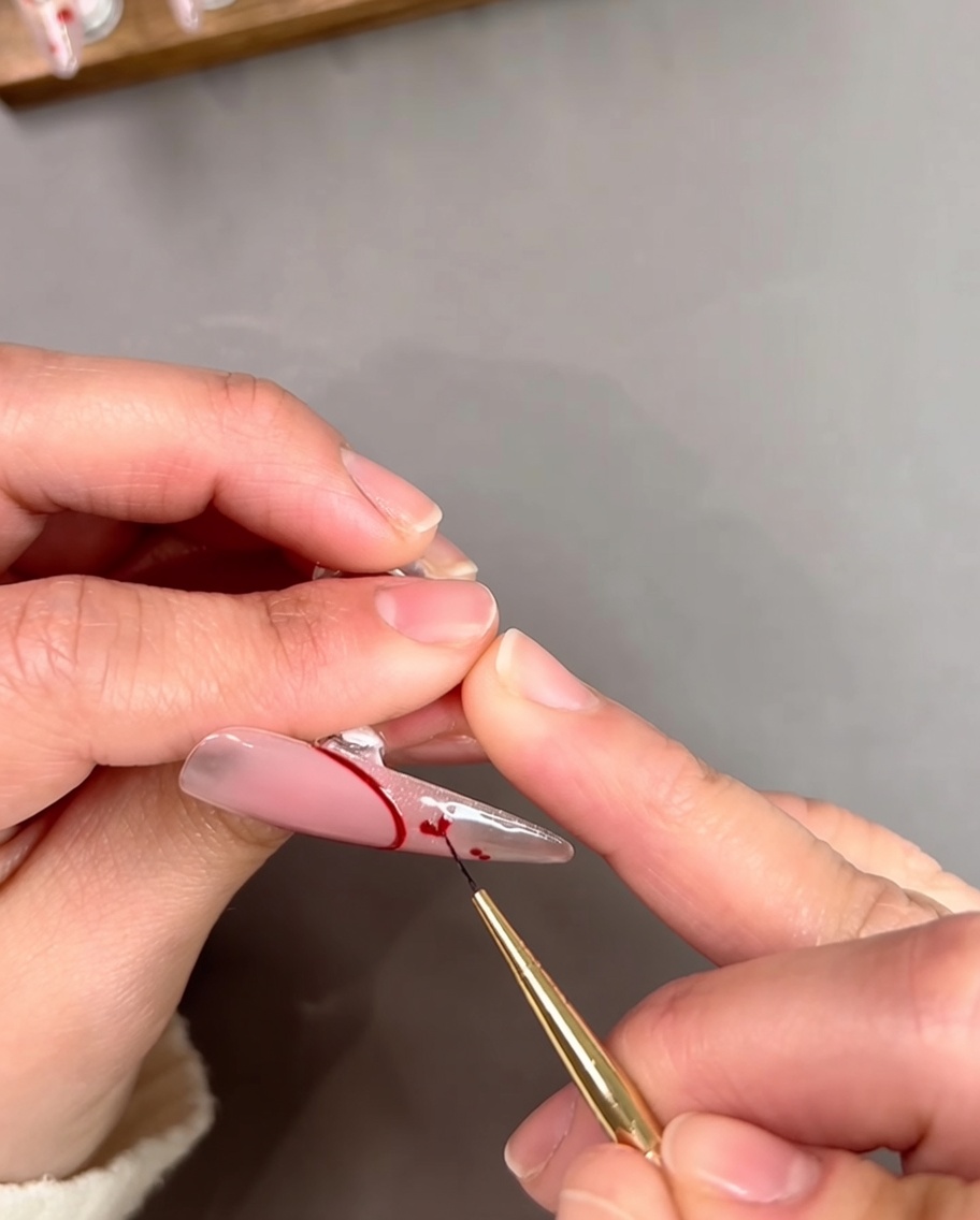 A close up shot of a nail artist using a thin brush to connect two small red dots into a heart shape on a clear nail tip.