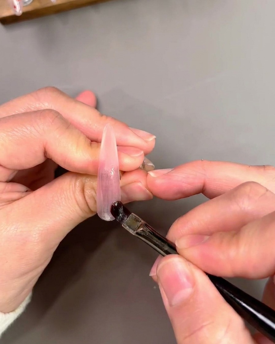 A flat gel brush applying and blending purple cat-eye gel polish onto an artificial nail tip.